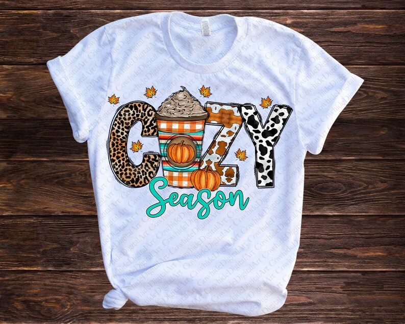 May include: White t-shirt with a graphic design of a coffee cup with whipped cream and a pumpkin, surrounded by animal print letters that spell out "Cozy Season".