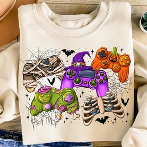May include: A cream-colored sweatshirt with a Halloween-themed design featuring four different video game controllers decorated with spooky elements like mummies, pumpkins, and skeletons. The controllers are surrounded by spiderwebs and bats.