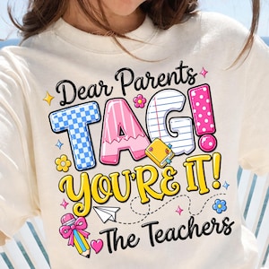 May include: Cream-colored sweatshirt with a colorful graphic that reads "Dear Parents TAG! YOU'RE IT! The Teachers." The design features playful fonts, a blue checkered "T", a pink "A", and a lined paper "G". Decorative elements include flowers, stars, and a pencil.