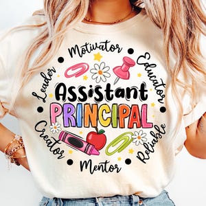 May include: Cream-colored t-shirt with a colorful graphic design. The design features the words "Assistant Principal" in large, multi-colored letters, surrounded by school-themed icons and words like "Leader" and "Educator".