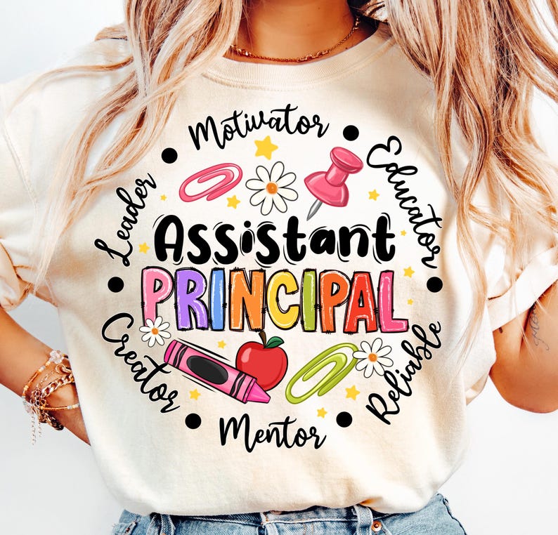 May include: Cream-colored t-shirt with a colorful graphic design. The design features the words "Assistant Principal" in large, multi-colored letters, surrounded by school-themed icons and words like "Leader" and "Educator".