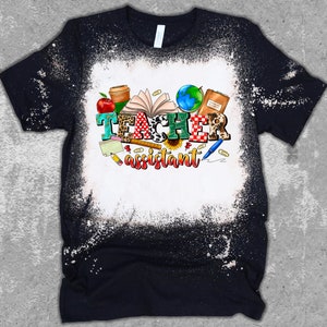K&ouml;nnte beinhalten: Ein schwarzes T-Shirt mit einem Bleiche-Spritzer-Design mit dem Text "Teacher assistant" umgeben von bunten Schulutensilien und einem Globus.
