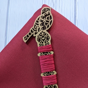 May include: A wooden bird-shaped thread holder with intricate cutouts and a red thread wound around it. The holder is on a red fabric background.