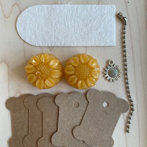 May include: A set of craft supplies including two yellow wax sunflower shapes, five brown paper spool-shaped tags, a white felt rectangle, and a silver charm with a sunflower design.
