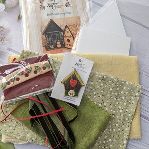 May include: A craft kit for making a birdhouse with a green and white floral fabric, green felt, red ribbon, a wooden birdhouse with a heart and the words "Love" on it, and a card with the words "Birdhouse in spring".