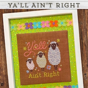May include: Framed cross-stitch art with the text "Ya'll Ain't Right." The artwork features three sheep with varying patterns and colors, stitched on brown fabric. The background is green with white polka dots, and the frame is white.