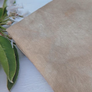May include: A close-up shot of a tan fabric with a mottled effect. The material is folded, showing its texture and colour variations. Green leaves and white flowers are in the background, adding a natural element to the composition.