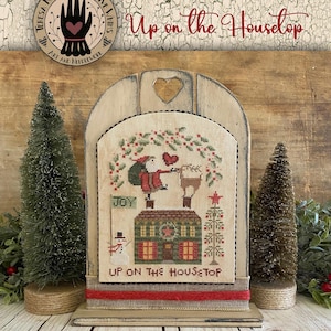 May include: A framed cross-stitch piece with the text "Up on the Housetop". The design features a Christmas scene with a house, snowman, and figures. The frame is light-colored wood with a heart cutout.