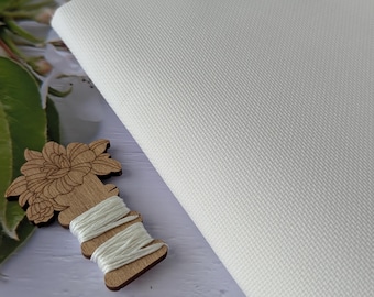 May include: A close-up of a white, textured fabric, possibly canvas, with a diagonal weave pattern. A wooden spool of white thread, a pair of pink handled scissors, and a wooden flower-shaped embellishment are also visible.