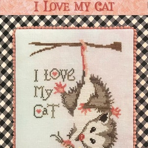 May include: Cross stitch pattern featuring a possum hanging upside down from a branch with the text "I love my cat" in pink and red hearts. The pattern is on a white background with a pink and black checkered border.