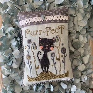 May include: An embroidered cushion featuring a black cat illustration and the text "Purr-fect". The cushion has a floral design, lace trim, and a small wooden heart. It is set against a backdrop of grey leaves.