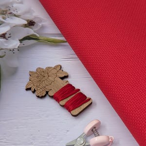 May include: A close-up of red cross-stitch fabric, wooden thread holder with red thread, pink scissors, and a pink and gold tassel. White flowers are in the background. The items are on a white wooden surface.