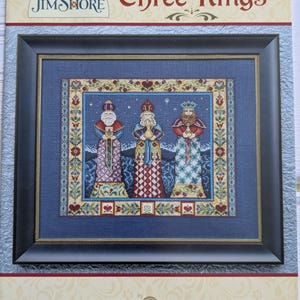May include: A framed cross-stitch pattern featuring three kings, each holding a gift. The artwork is set against a blue background with a decorative border. The text "Three Kings" is displayed at the top, with the Jim Shore and Mill Hill logos.