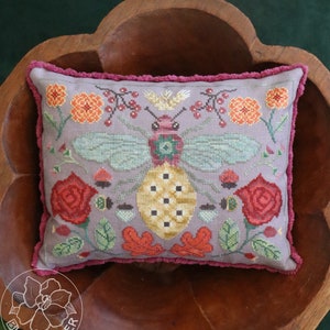 May include: A decorative cushion with a hand-stitched design featuring a bee, flowers, and leaves. The cushion is made of a soft, light purple fabric with a pink velvet trim.