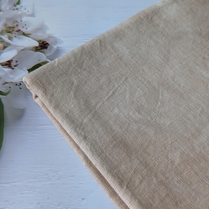 May include: Folded beige linen fabric, displaying a natural, textured weave. The fabric has a subtle, earthy tone, with slight colour variations. The image is lit with soft, natural light, highlighting the fabric's texture and drape. White flowers are in the background.