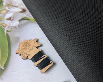 May include: A black, textured fabric is the focal point of this image. A wooden spool of black thread, a pair of small scissors with pink handles, and a decorative tassel are also present. The items are arranged on a white wooden surface.