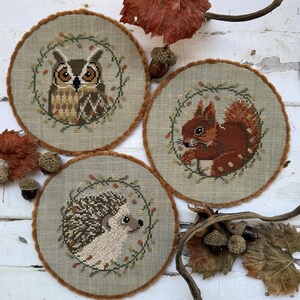 May include: Three circular cross-stitch designs showcasing woodland animals. An owl, squirrel, and hedgehog are depicted on a light green fabric, encircled by a wreath of leaves and berries. Each design has a brown, fuzzy border.