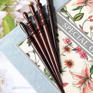 May include: Five dark brown wooden hair sticks with decorative tops are arranged on a floral patterned background. The sticks vary in design, with some having carved details. The background includes the text "SPECIAL G".