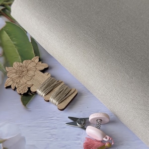 May include: A neutral-toned fabric, likely linen or cotton, is partially visible, alongside cross-stitch supplies. A wooden spool of thread, small scissors, and a pink tassel with gold accents are also present, suggesting a crafting theme.