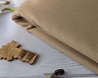 May include: A beige fabric with a textured weave. The fabric is folded in half and lying on a white wooden surface. There are also a wooden thread holder with a floral design, a pair of small scissors with a pink tassel, and a spool of thread.