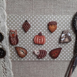 May include: Nine hand-painted ceramic buttons featuring autumnal themes. The buttons depict an owl, a pumpkin, an acorn, a hedgehog, two leaves, and a squirrel.