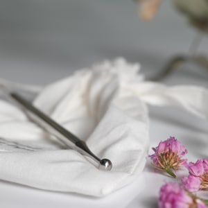 May include: A silver metal tool with a rounded tip, resting on a white fabric pouch.  Pink flowers are in the background.