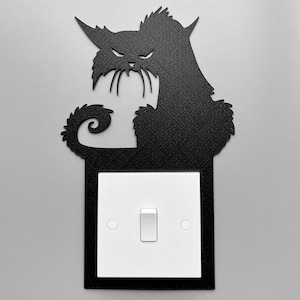 May include: Black metal light switch cover in the shape of a cat. The cat has a grumpy expression, pointed ears, and a curled tail. The cover surrounds a white light switch with a rectangular toggle.