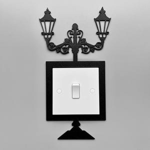 May include: Black metal light switch cover featuring a decorative street lamp design. The cover surrounds a white light switch with a single toggle. The design includes two lamp fixtures and ornate scrollwork.