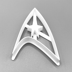Puede incluir: Cortador de galletas de pl&aacute;stico blanco con forma del emblema de Star Trek. El cortador tiene una gran estrella de cinco puntas en el centro.