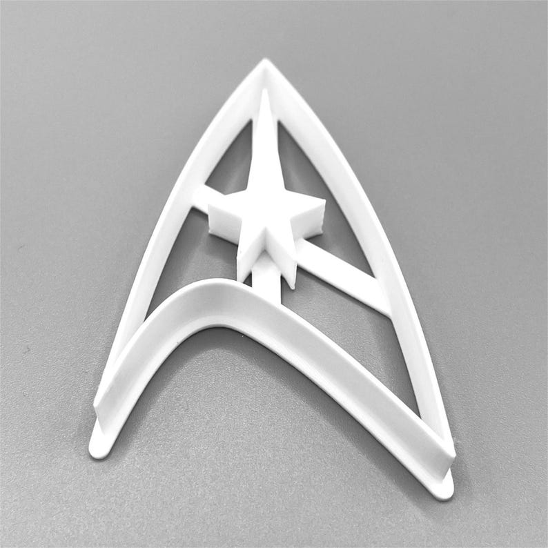 Puede incluir: Cortador de galletas de pl&aacute;stico blanco con forma del emblema de Star Trek. El cortador tiene una gran estrella de cinco puntas en el centro.