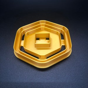 May include: A gold-coloured plastic cookie cutter shaped like a hexagon with three concentric rings. The cutter has a square opening in the centre.