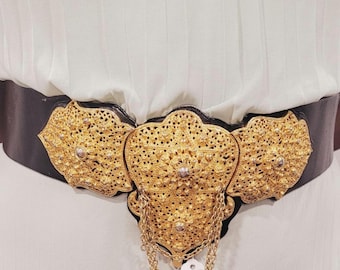 Könnte beinhalten: Ein schwarzer Gürtel mit aufwendigen goldfarbenen Metallarbeiten. Der Gürtel hat ein zentrales Dekorationselement mit einer hängenden Kette und zwei Seitenteilen. Die Metallarbeiten haben ein detailliertes, offenes Design.
