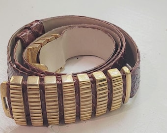 May include: A brown belt with a textured snakeskin pattern. The belt features a series of rectangular gold-colored metal links as a buckle. The belt is rolled up, showing the interior lining.
