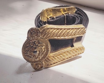 Könnte beinhalten: Ein schwarzer Gürtel mit einer goldfarbenen Schnalle. Die Schnalle hat ein verziertes Design mit einem kreisförmigen Emblem und einem rechteckigen Rahmen. Der Gürtel besteht aus einem strukturierten Material.