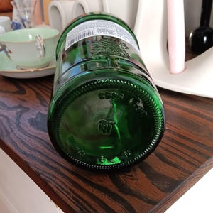 May include: A green glass bottle with a textured base and a label around the top. The bottle is lying on a dark wooden surface. The base of the bottle has embossed markings.