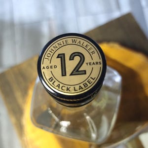 May include: A clear glass bottle with a black and gold cap. The cap reads "JOHNNIE WALKER" and "BLACK LABEL 12 YEARS AGED". The bottle's square shape is partially visible.