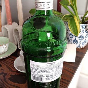 May include: A green glass bottle of Tanqueray London Dry Gin with a silver cap. The bottle has embossed lettering and a label with nutritional information. The bottle is on a wooden surface.