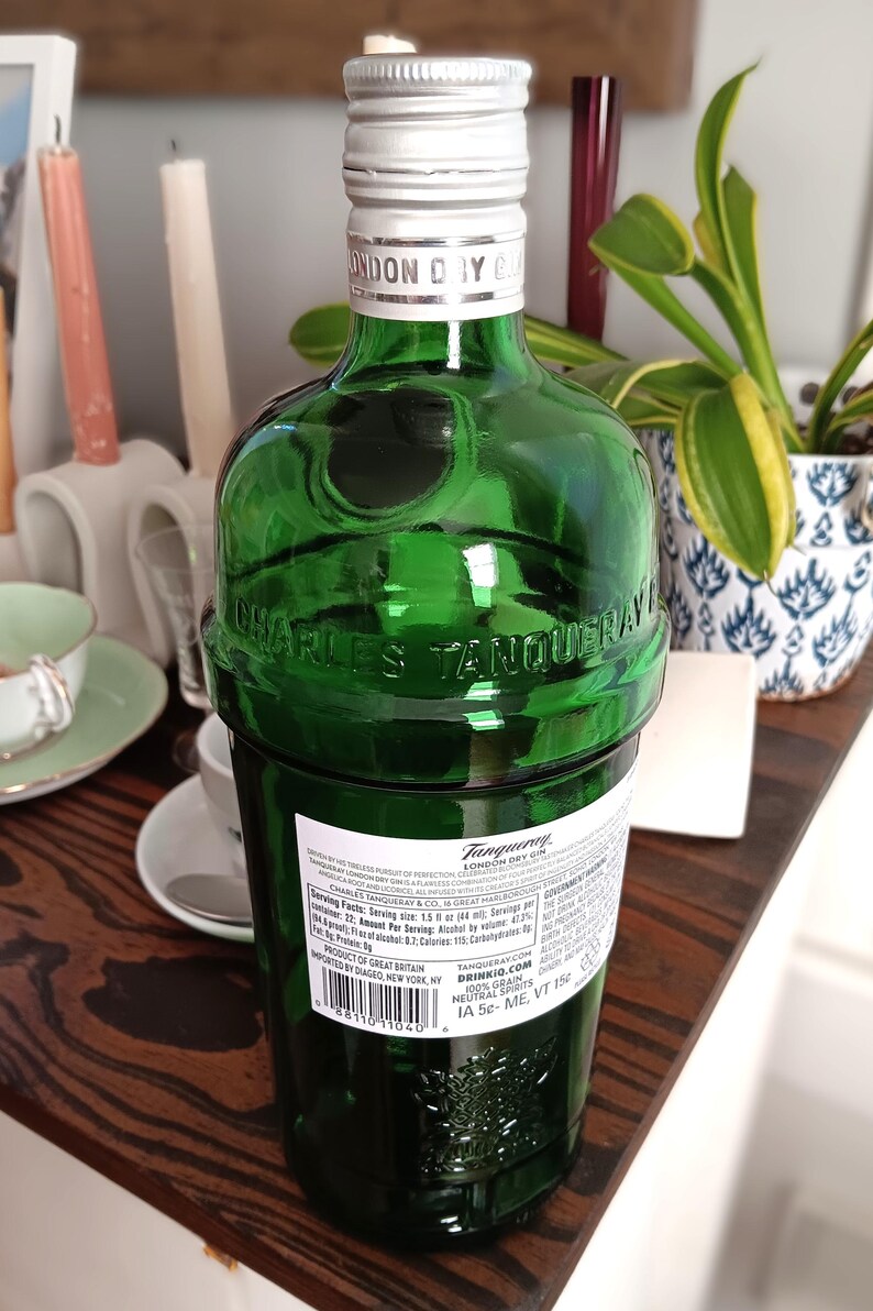 May include: A green glass bottle of Tanqueray London Dry Gin with a silver cap. The bottle has embossed lettering and a label with nutritional information. The bottle is on a wooden surface.