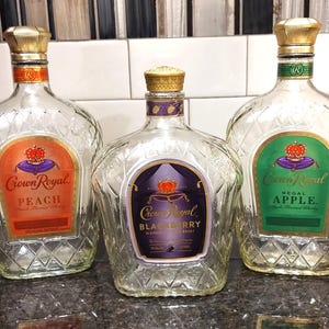 May include: Three clear glass bottles of Crown Royal flavored whiskey. The bottles are arranged in a row from left to right: Peach, Blackberry, and Regal Apple. Each bottle has a gold cap and a diamond-shaped pattern.