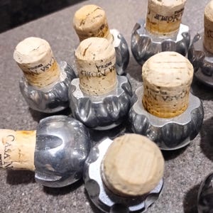 May include: Close-up of nine silver bottle stoppers with cork tops. The corks are labeled with the text "CASA NOVA" and "JOMBA".
