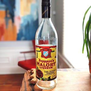 May include: A clear glass bottle of Jeppson's Malört Liqueur with a black cap. The label is yellow with red and blue accents, and the text "Jeppson's Malört Liqueur" is prominently displayed. The bottle is on a wooden surface.
