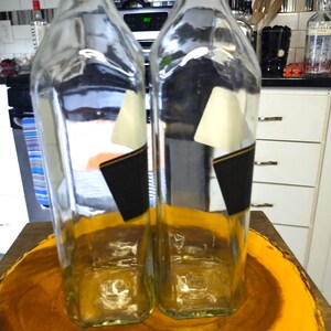 May include: Two clear glass bottles with black and white labels. The bottles are rectangular with rounded shoulders and a square base. The bottles are sitting on a wooden surface.