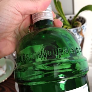 May include: A green glass bottle of Tanqueray London Dry Gin. The bottle has embossed lettering and a silver cap. The label is white with black text. The bottle is held by a hand.