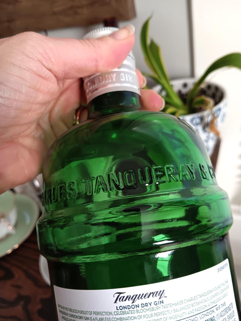 May include: A green glass bottle of Tanqueray London Dry Gin. The bottle has embossed lettering and a silver cap. The label is white with black text. The bottle is held by a hand.