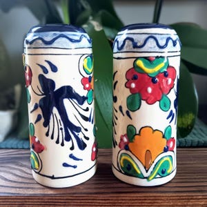 May include: Two cylindrical ceramic salt and pepper shakers featuring a cream-colored base with hand-painted designs. The shakers are decorated with dark blue bird motifs, vibrant red, orange, and green floral patterns, and blue wavy line borders.