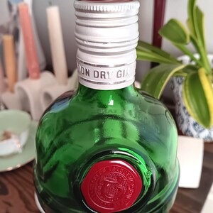 May include: A green glass bottle of London Dry Gin with a silver cap and a red wax seal. The bottle has a rounded shape and the words "LONDON DRY GIN" are printed on the cap. The seal features an emblem and text.