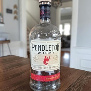 May include: A clear glass Pendleton Whisky bottle with a wooden stopper. The label features the brand name, a red image of a horse and rider, and the text "True Western Tradition". The bottle is on a wooden surface.