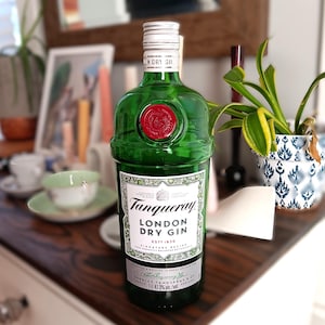 May include: A green Tanqueray London Dry Gin bottle with a red wax seal. The label reads "Tanqueray LONDON DRY GIN ESTD 1830". The bottle is 11.47.3% alc/vol. The bottle is on a dark wooden surface.