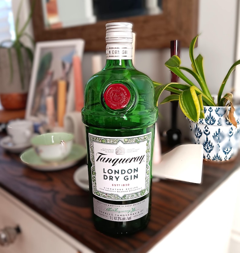 May include: A green Tanqueray London Dry Gin bottle with a red wax seal. The label reads "Tanqueray LONDON DRY GIN ESTD 1830". The bottle is 11.47.3% alc/vol. The bottle is on a dark wooden surface.