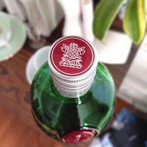 May include: A green glass bottle with a silver cap and a red top. The red top features a white pineapple design. The bottle is partially visible, showcasing its rounded shape and a red seal at the base.